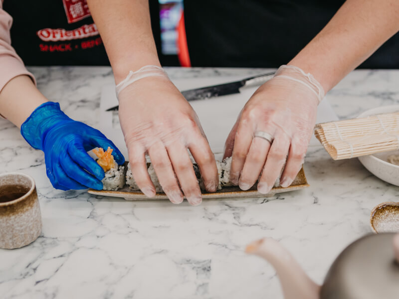 A close up of two pairs of hands wearing gloves positioning their handmade sushi.